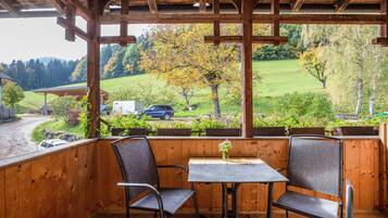 Un espace extérieur abrité, aménagé avec deux chaises et une table, qui offre une vue sur un paysage verdoyant, avec des arbres et une voiture garée.