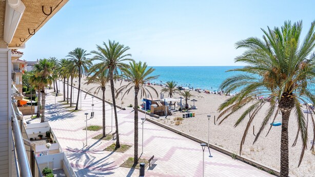 A beach view with palm trees, a clear sky, and a building with balconies. - Coral Beach by Pat Apartment with Sea Views, Shared Pool, and Wi-Fi (Calafell)