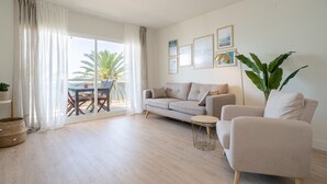 A living room with a sofa, armchair, and a small round table. There is a balcony with a table and chairs, and a large plant. - Coral Beach by Pat Apartment with Sea Views, Shared Pool, and Wi-Fi (Calafell)
