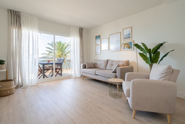 A living room with a sofa, armchair, and a small round table. There is a balcony with a table and chairs, and a large plant.