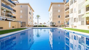 A swimming pool surrounded by multi-story buildings with balconies, a palm tree, and outdoor umbrellas. - Coral Beach by Pat Apartment with Sea Views, Shared Pool, and Wi-Fi (Calafell)