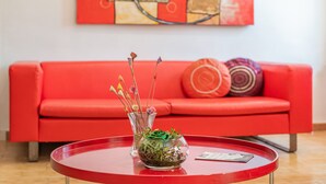 Un sofá rojo con un florero de vidrio que contiene plantas verdes y ramas decorativas, una mesa roja con una tarjeta, y una pared roja con arte abstracto.