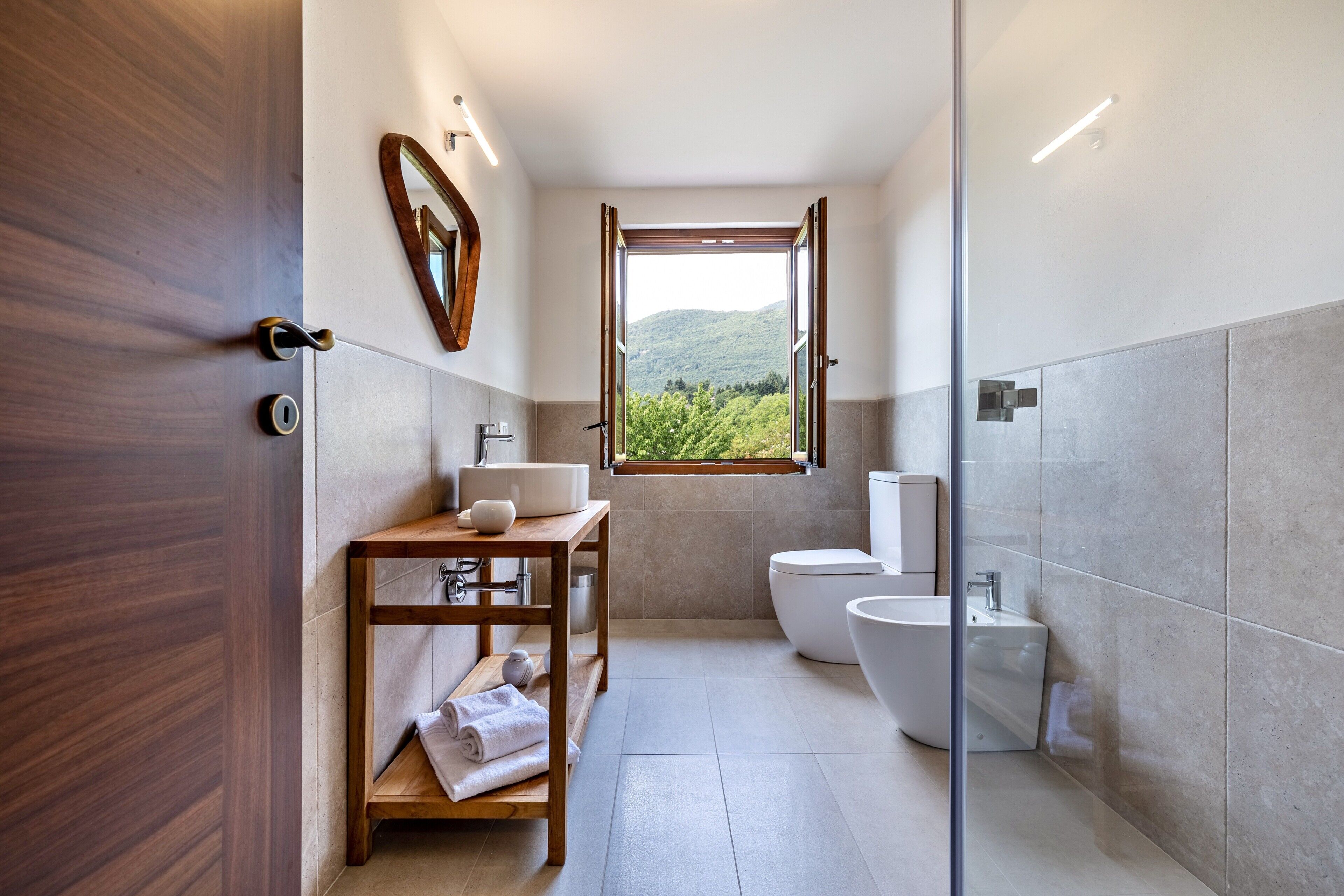 A modern bathroom with a wooden vanity, a toilet, a bidet, and a window with a view of greenery.