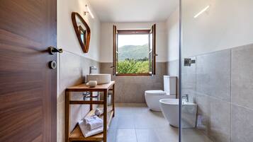 A modern bathroom with a wooden vanity, a toilet, a bidet, and a window with a view of greenery.