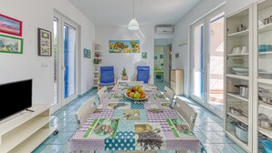 A dining area with a table set for four, a television on the left, and a kitchen area with open shelving to the right.