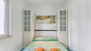 A small, single-bed room with a colorful bedspread, a painting above the bed, and two towels on the bed.