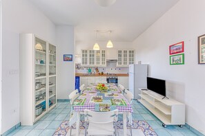 A compact kitchen with white cabinets, a dining table with chairs, a TV on a stand, and a patterned rug.