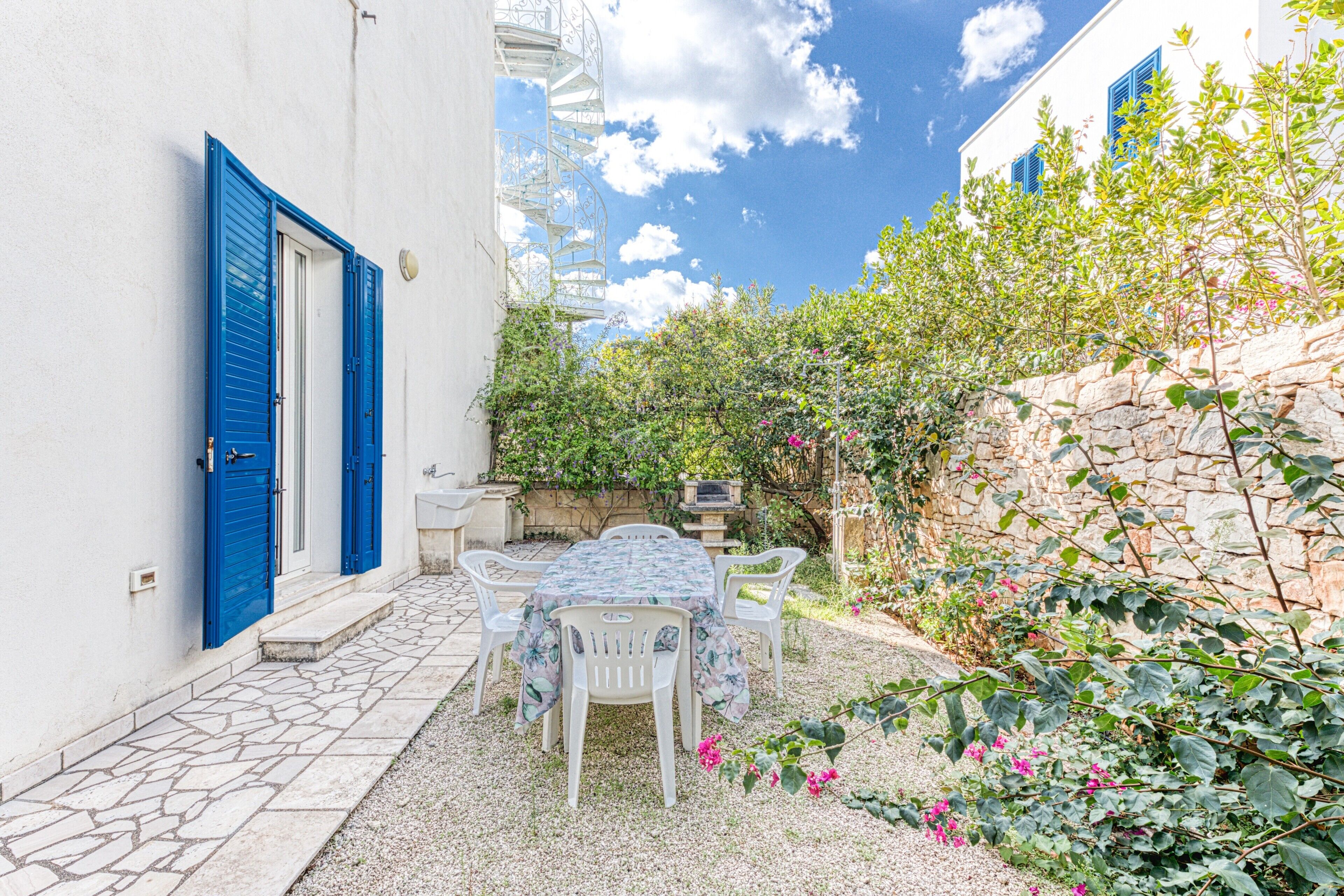 Un cortile con tavolo e sedie, un edificio bianco con persiane blu e un muro in pietra.