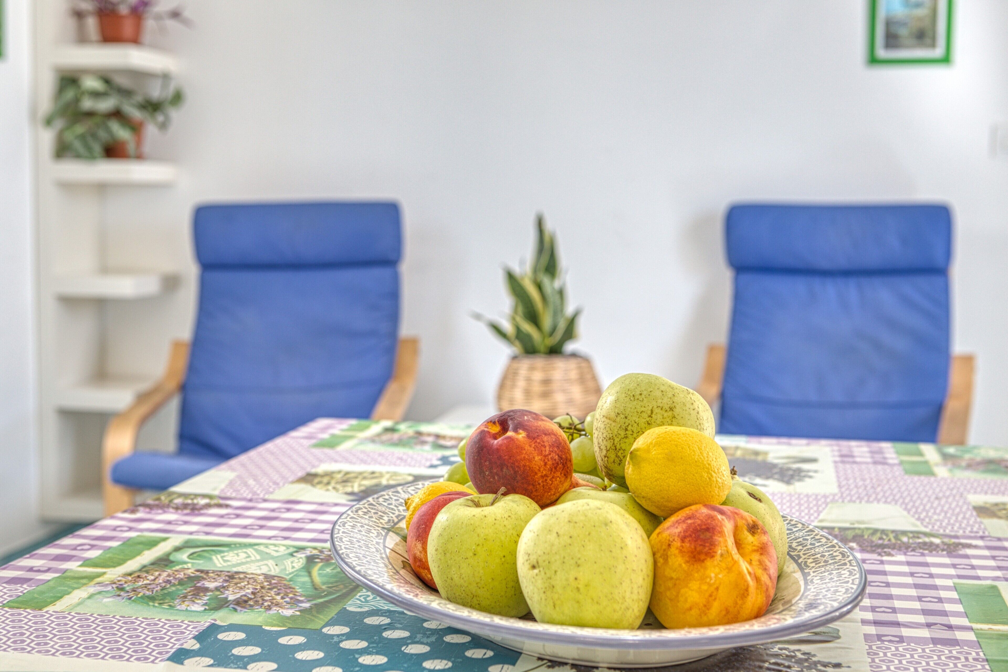 Een eettafel met een fruitschaal, twee blauwe stoelen, een potplant en een tafelkleed met dessin.