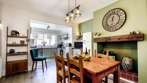A dining area with a wooden table and chairs, a green wall with a clock, and a kitchen in the background.