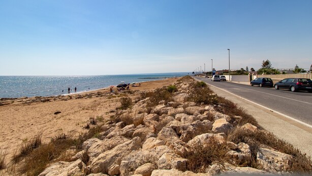 Una carretera costera con playa, terreno rocoso y coches aparcados.