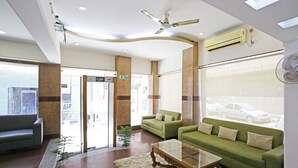 A reception area with a green sofa, a glass coffee table, and a white rug. There is a ceiling fan, an air conditioning unit, and a view of cars outside. - MyHomeHotels – Hotel Ambrosia (New Delhi)