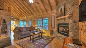 Interior acogedor de una cabaña de troncos con chimenea de piedra, muebles de madera y vistas al exterior a través de amplias ventanas.