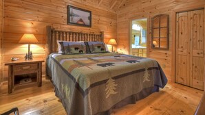 Un dormitorio de cabaña de madera con una cama, dos mesitas de noche con lámparas, una ventana y una puerta.