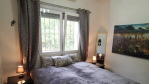 A bedroom with a bed, two bedside lamps, a window with curtains, a wall-mounted mirror, and a cityscape painting.