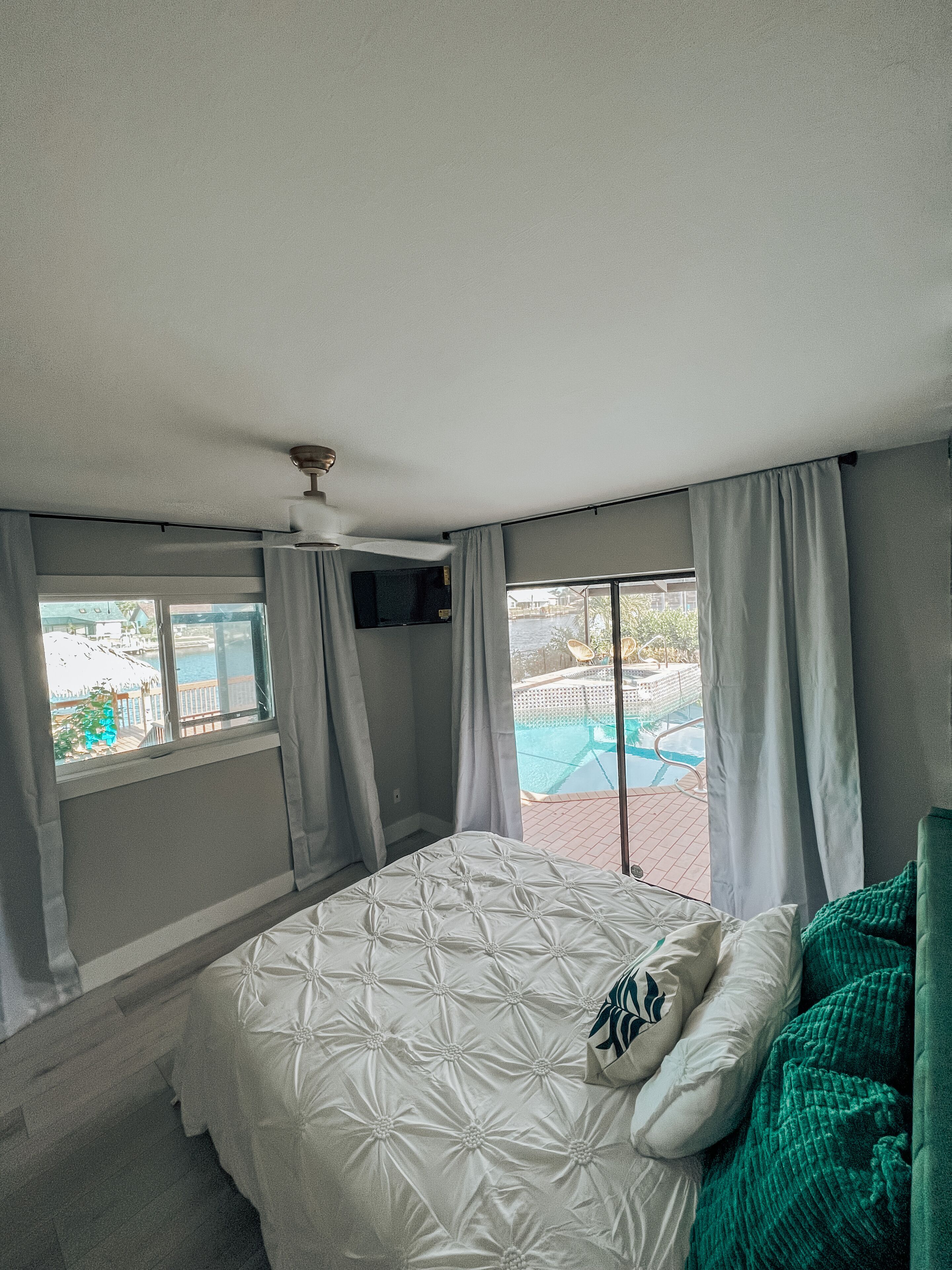 A bedroom with a bed, a window with curtains, a ceiling fan, and a view of an outdoor swimming pool.