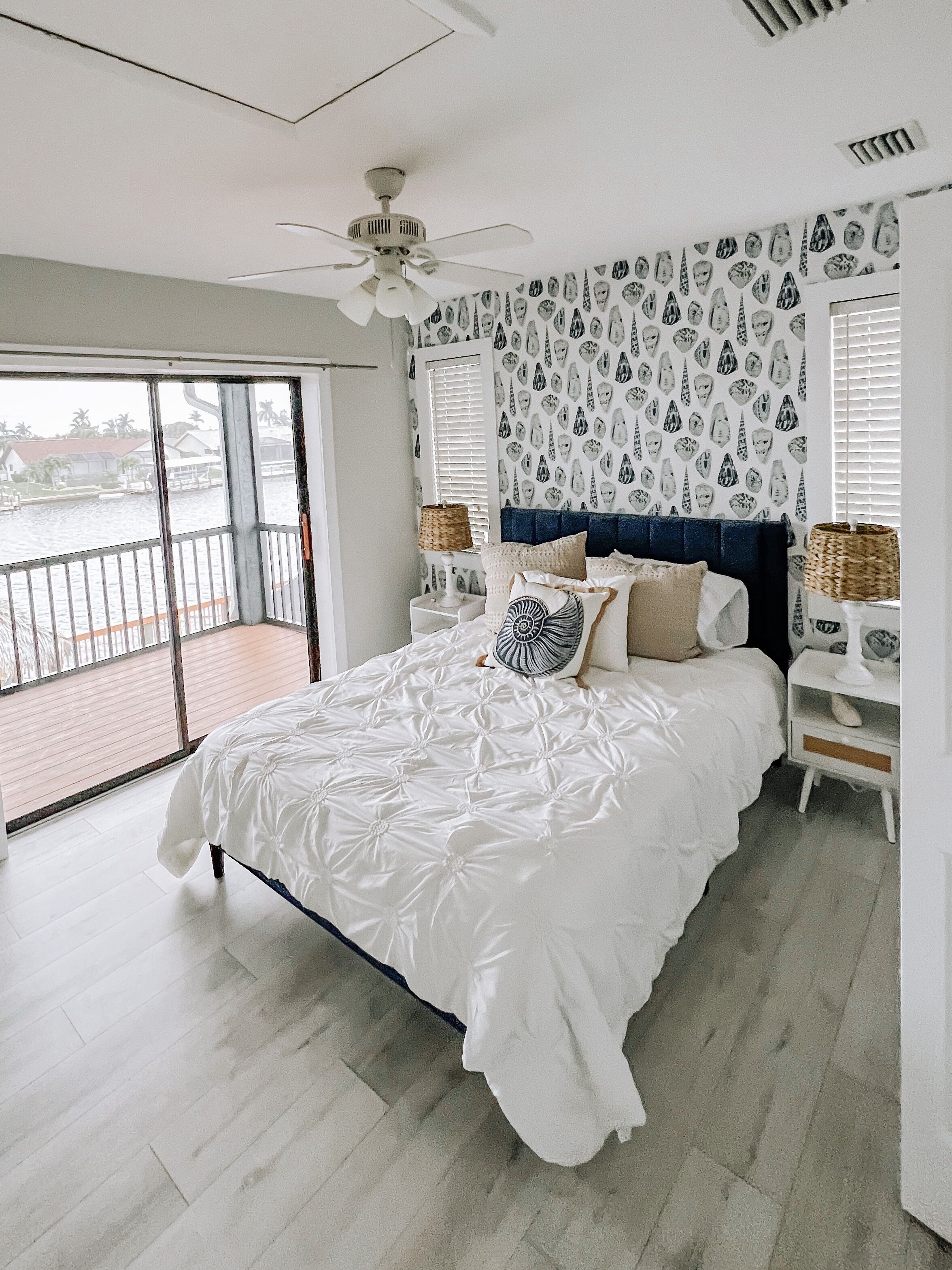 A bedroom with a large bed, a ceiling fan, a patterned wall, and a view of the water outside.