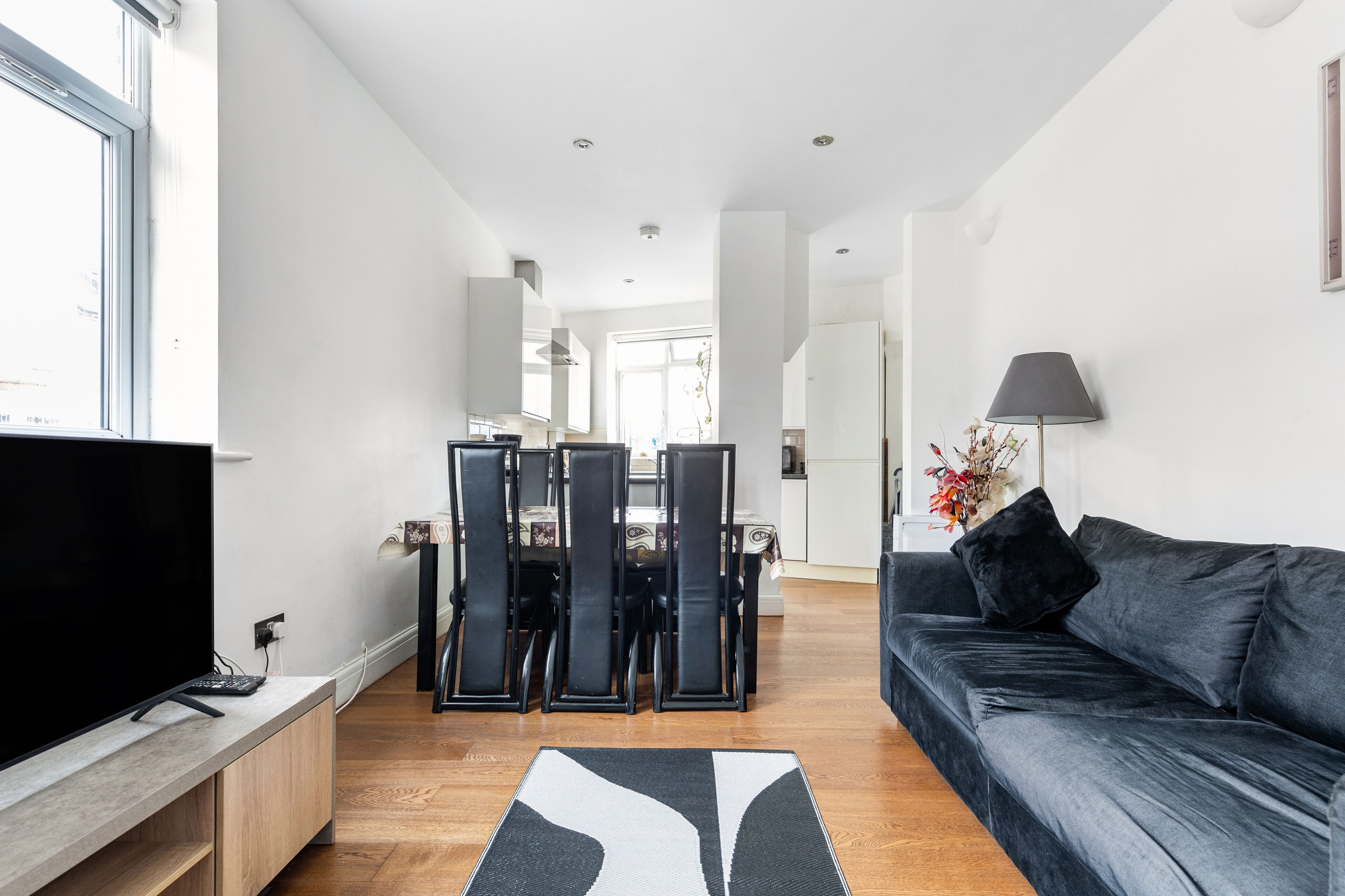A modern living room with a flat-screen TV, a dark sofa, and a dining area with chairs and a table.