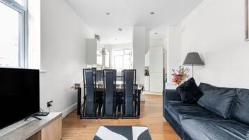 A modern living room with a flat-screen TV, a dark sofa, and a dining area with chairs and a table.