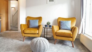 Een woonkamer met twee gele fauteuils, een rond geweven zitkussen, een kleine tafel met een plant en een lijst met een afbeelding aan de muur.