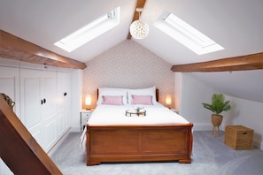 Une chambre avec un grand lit, des fenêtres de toit, des poutres en bois, une table de chevet, des lampes de chevet, une plante en pot et un panier en osier.