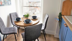 Een eetruimte met een ronde houten tafel, vier grijze gestoffeerde stoelen, en een uitzicht op de stad door een groot raam.