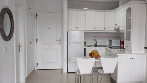 Une cuisine avec des meubles blancs, un réfrigérateur, un four à micro-ondes et un évier. On y trouve deux chaises blanches et une petite table avec des fruits.
