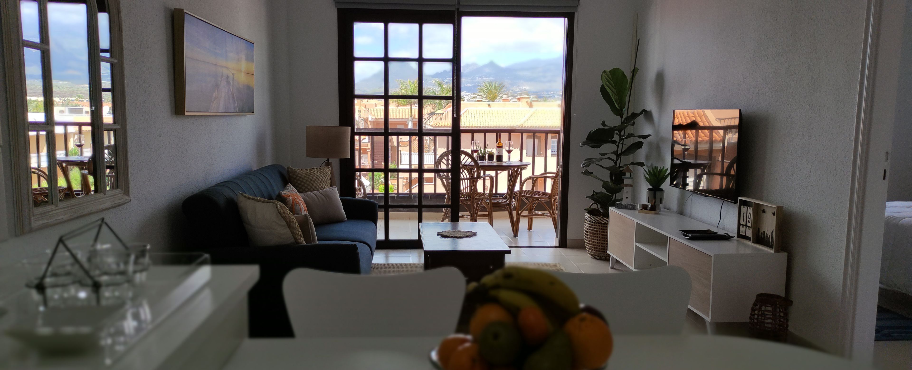 A living room with a sofa, coffee table, and a bowl of fruit. There is a balcony with a table and chairs.