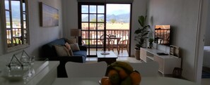 Uma sala de estar com sofá, mesa de centro e uma tigela de frutas. Há uma varanda com mesa e cadeiras.