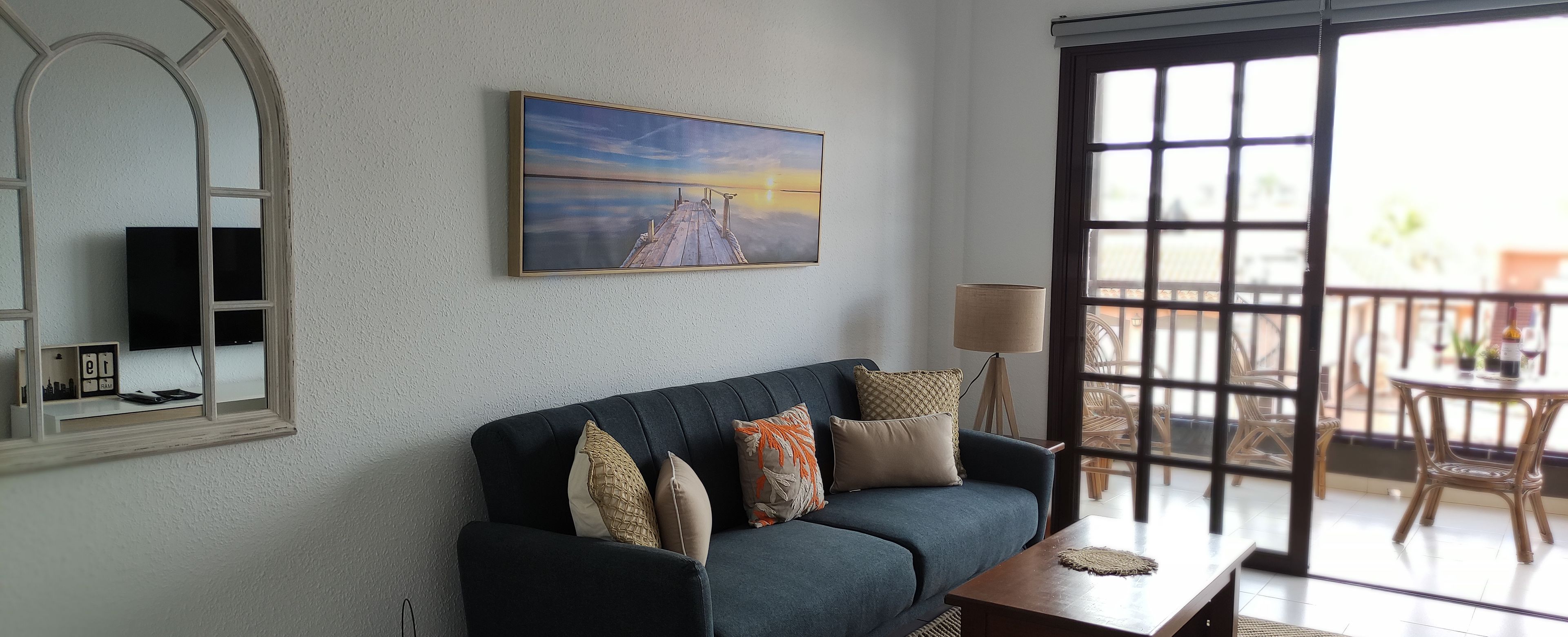 Een woonkamer met een donkerblauwe bank, een schilderij van een zonsondergang, een televisie en een balkon met rieten meubels.