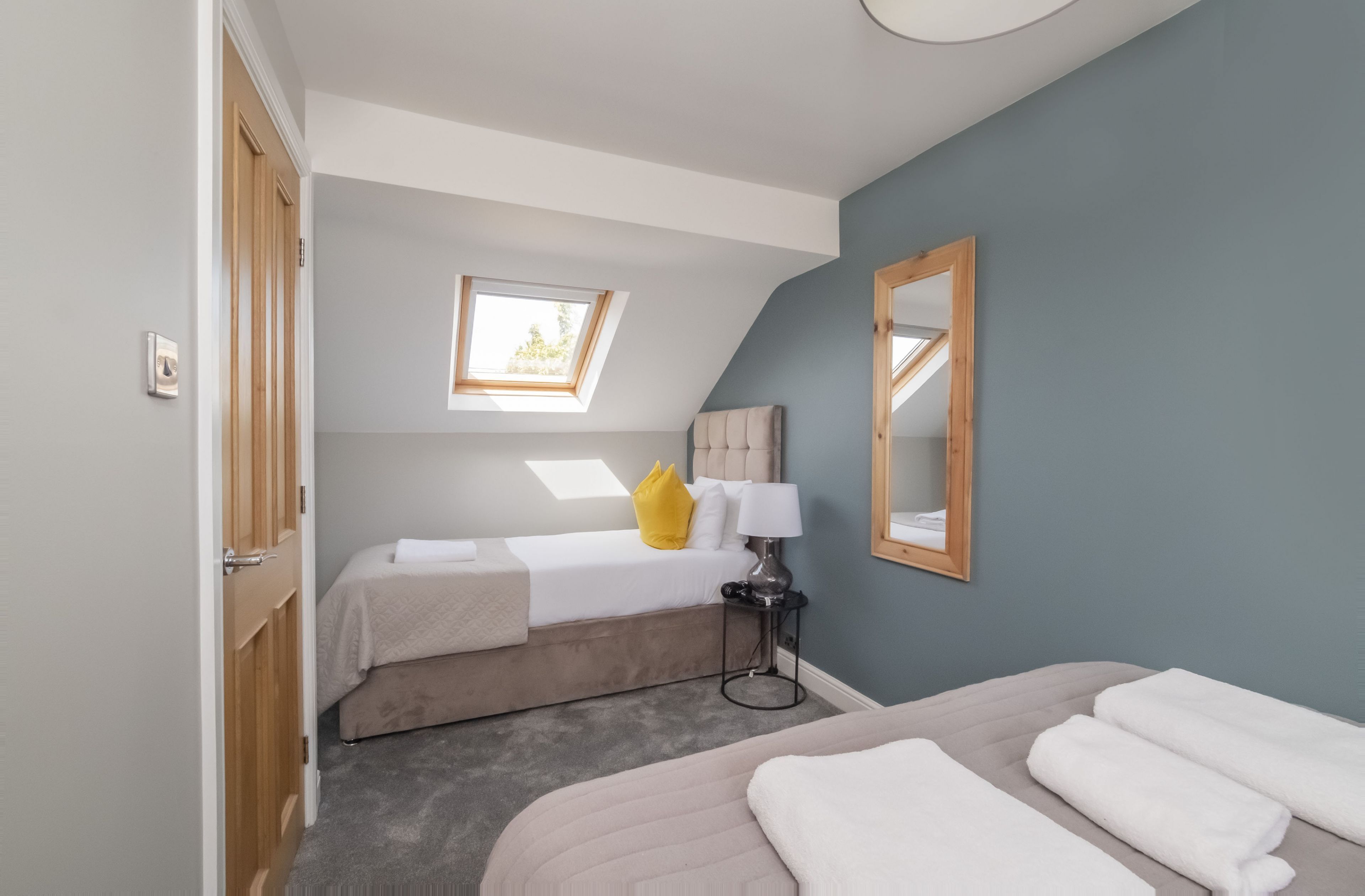A bedroom with a bed, a bedside table with a lamp, a window, and a mirror on the wall.