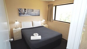 Una cama bien hecha con una colcha oscura y almohadas blancas, una lámpara de noche, una mesita con lámpara, y un cuadro en la pared.