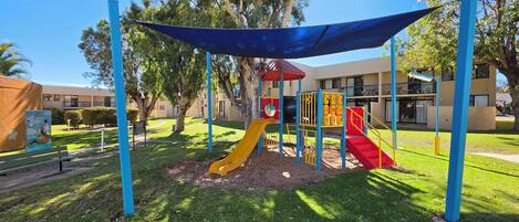 A playground with slides and climbing structures, surrounded by trees and a building in the background.