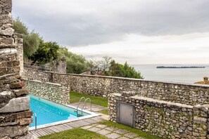 Une piscine en pierre offrant une vue sur la mer, entourée de murs de pierre et d’une zone herbeuse.