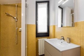 Une salle de bain avec des murs recouverts de carreaux jaunes, un lavabo blanc, un miroir et une douche équipée d’un pommeau de douche à main.