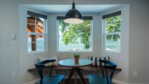 Een eethoek met een ronde tafel, vier stoelen, een hanglamp en drie ramen met uitzicht op groen.