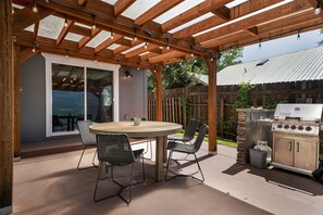 Un patio al aire libre con una mesa redonda y sillas, una parrilla y una pérgola con luces colgantes.