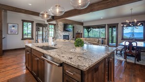 A spacious kitchen with a granite countertop, stainless steel appliances, and a stone fireplace. There is a dining area with a wooden table and chairs, and large windows offering views of the outdoors.