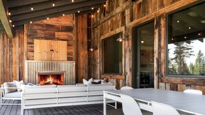 A wooden deck with a fireplace, white furniture, and large windows overlooking a forest.