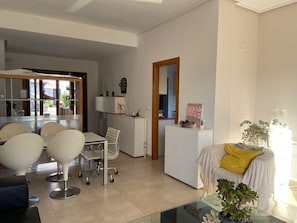 A modern living room with a glass coffee table, a sofa with a yellow throw, and a dining area with a white table and chairs.