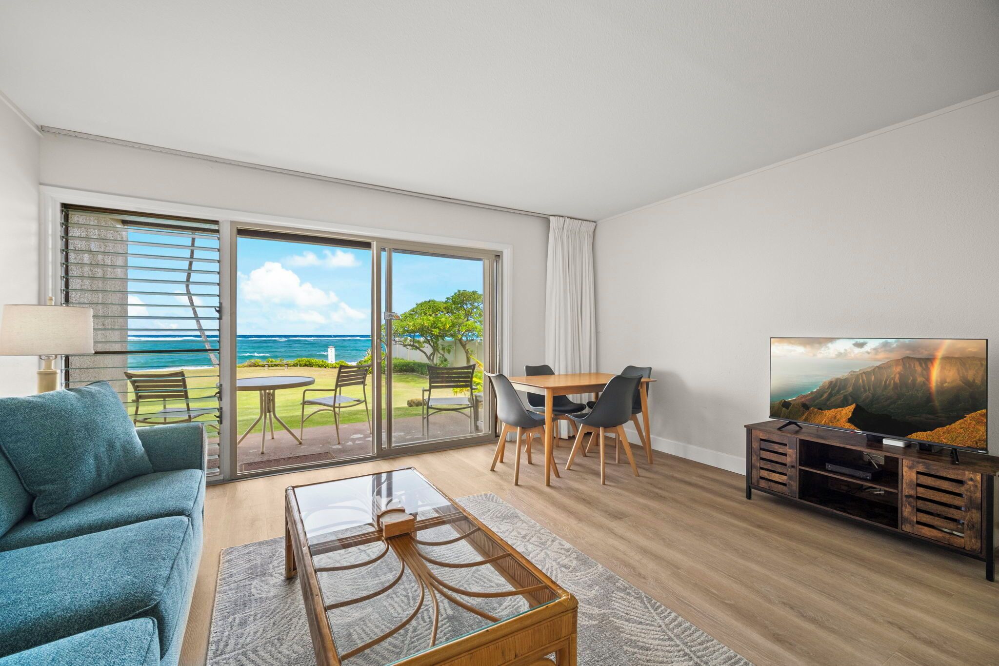 A modern living room with a large sliding glass door leading to a balcony with a view of the ocean. There is a sofa, a coffee table, and a TV on a wooden stand.