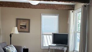 Una sala de estar con un sofá gris, una mesa de centro blanca, un televisor sobre un mueble de madera y una ventana con cortinas.