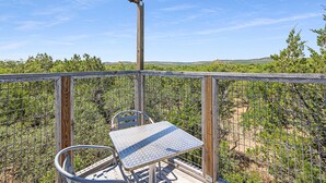 A balcony with a metal table and chair, overlooking a fenced garden with trees and a clear sky. - Come On Inn | Close to Downtown Wimberley! (Wimberley)