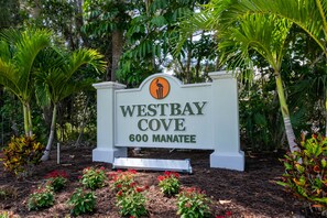 A sign for Westbay Cove, 600 Manatee, surrounded by lush greenery and flowering plants.
