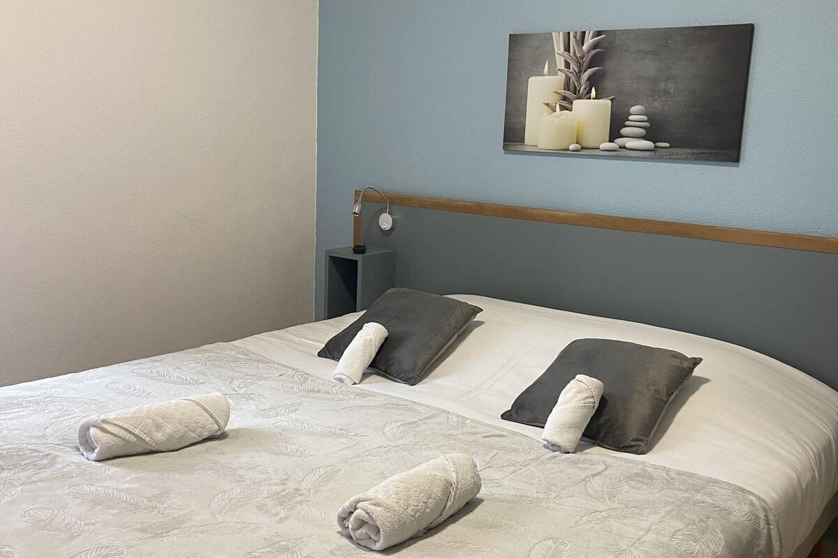 Una cama bien tendida con una colcha de estampado de hojas, dos almohadas y dos toallas enrolladas.