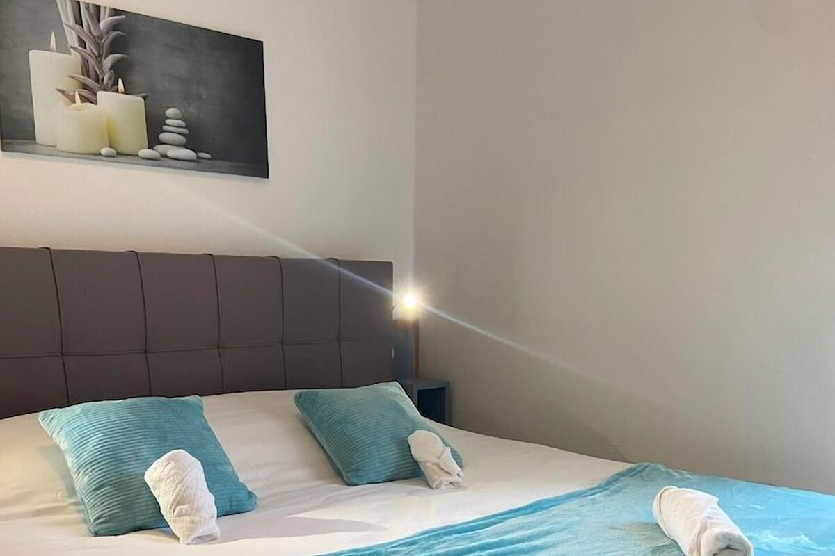 Una cama bien hecha con una colcha verde azulada, dos almohadas y toallas enrolladas.