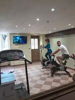 A fitness room with stationary bikes, a treadmill, and a TV displaying a movie.