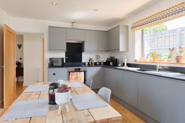 A modern kitchen with grey cabinets, a black stove, and a window with a wooden blind. - Seadog (Porthleven)