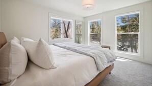 Ein Schlafzimmer mit einem großen Bett, weißer Bettwäsche und mehreren Fenstern mit Blick auf Bäume und Himmel.