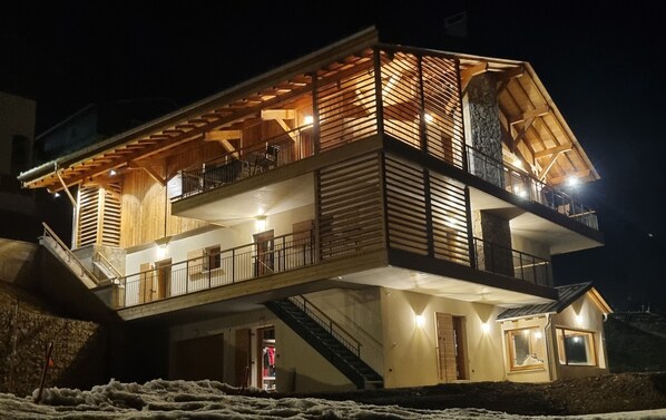 A multi-story wooden building with balconies and staircases, featuring a stone facade and a warm interior lighting.